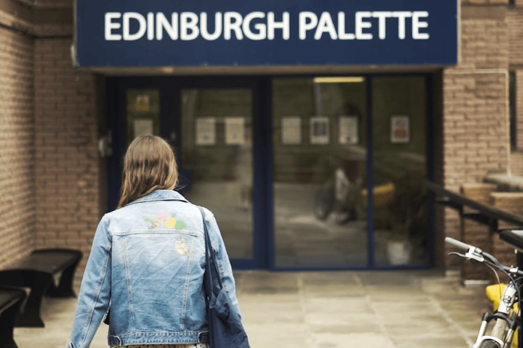 A person wearing a denim jacket walking into an entrance of a building with a sign that reads "Edinburgh Palette.