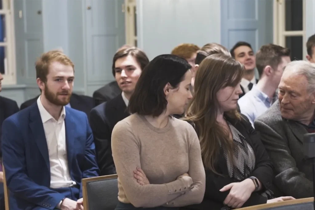 A diverse group of people seated in a formal setting, engaged in conversation during an event, showcasing various expressions and interactions.
