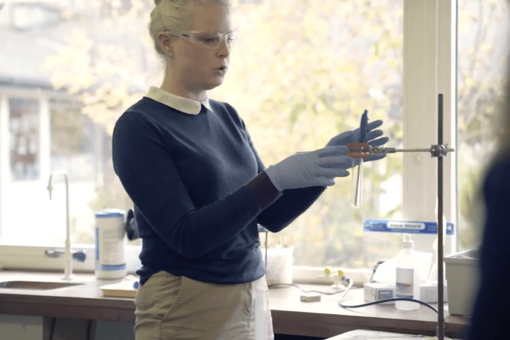 Teacher with light blonde hair and white shirt under dark blue sweater puts on blue protective gloves and stands in front of a window in a laboratory room with laboratory equipment on the work surface