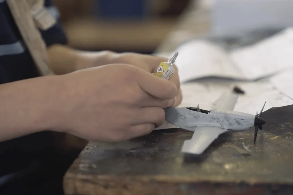 Hand holding an adhesive container over a grey model aircraft on a table