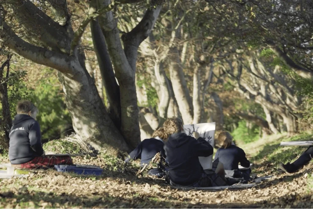 Group of children sitting on the floor under trees outdoors, some wearing dark tops