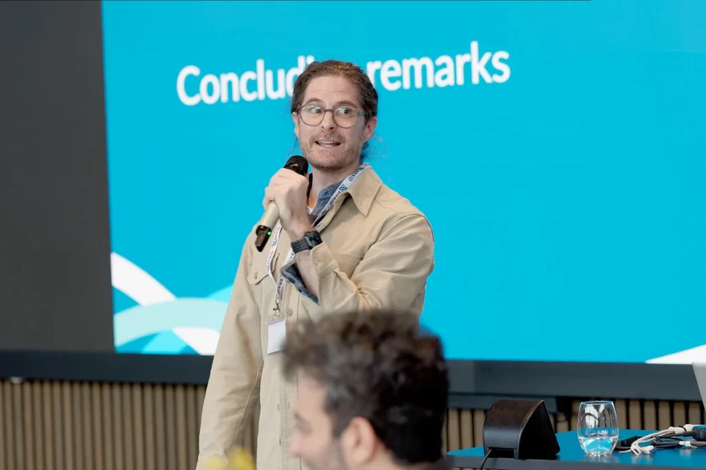 Person with microphone in front of large screen with text 'Concluding remarks' at an event