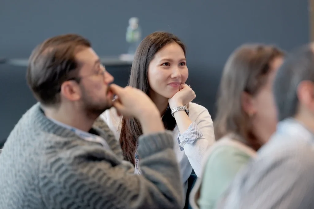 Group of people in a seminar room looking intently forward, one person wearing a grey cardigan, another a white blouse with a wristwatch