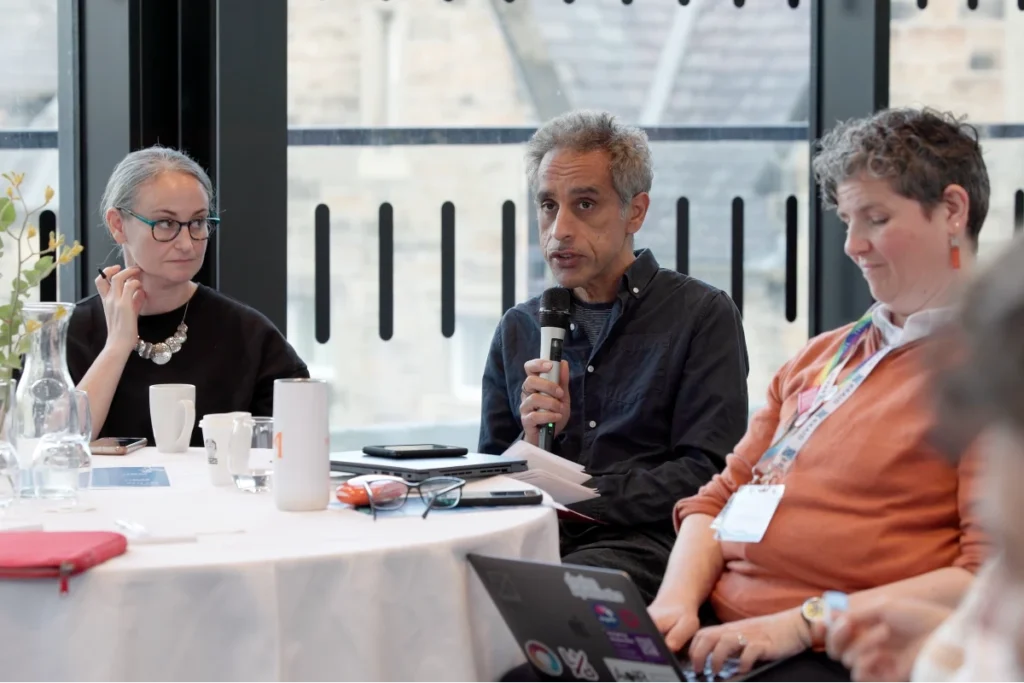 Three people sit at a round table with white tablecloth, one person holds a microphone, on the table are cups, a water bottle and a laptop with stickers.