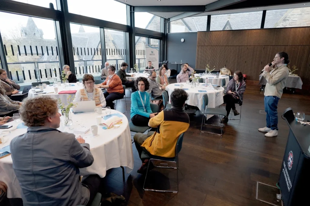 Several people sit at round tables in a modern, light-flooded room and listen to a standing person with a microphone.