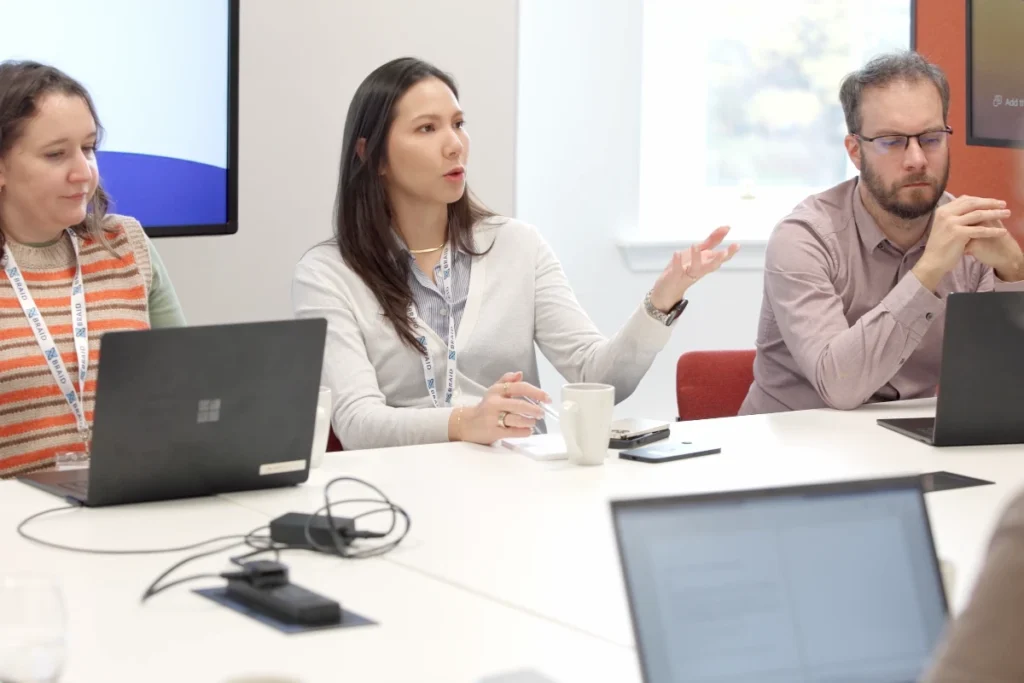 Three people sit at a table with laptops and discuss in a bright meeting room.