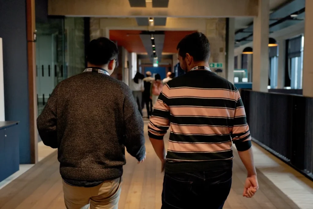Two men with name tags walk away from the camera in a modern hallway with wooden floor and large windows