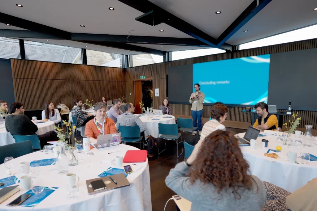 Several people sit at round tables with white tablecloths in a modern conference room, while a man speaks in front of a large blue presentation wall.