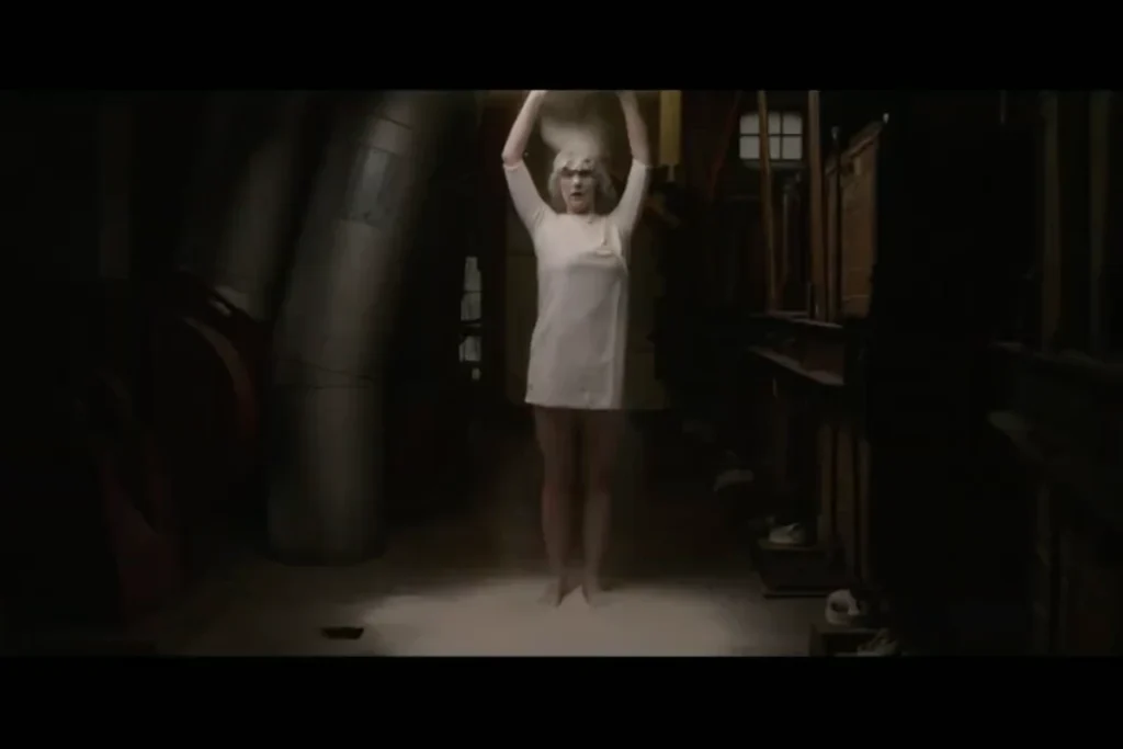 A woman in a white dress stands in a dimly lit room, raising her arms in a dramatic pose, surrounded by industrial elements.