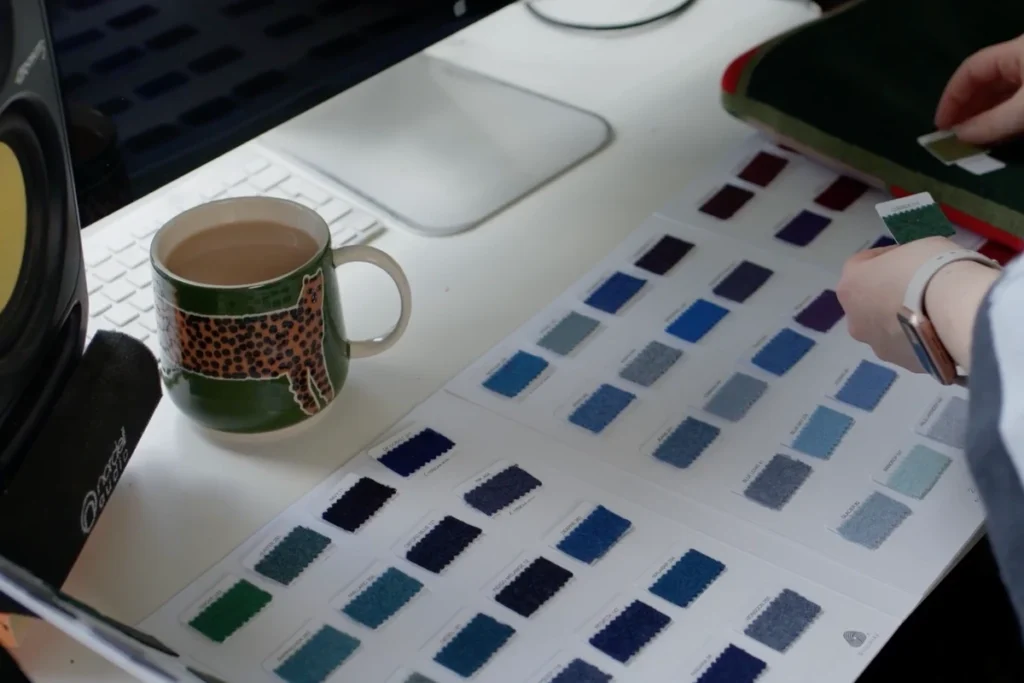 Hand holds color pattern card over a table with swatches in different shades of blue and green, next to cup with leopard pattern and green stripe