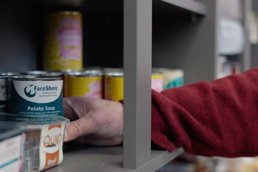 A hand reaching for a can of FareShare potato soup on a shelf, promoting food security and reducing waste.