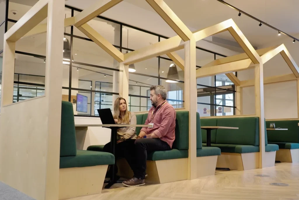 Two professionals engaged in a discussion at a modern workspace with cosy seating and a laptop on the table.