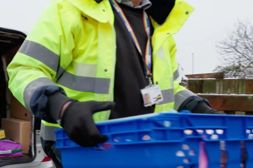 A delivery worker in a bright yellow jacket holds a blue crate while unloading items from a van in a snowy environment.