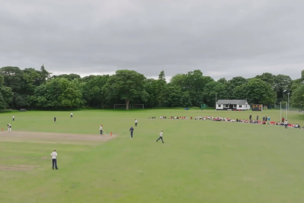 A cricket match in progress on a lush green field with spectators seated in the background under a cloudy sky.