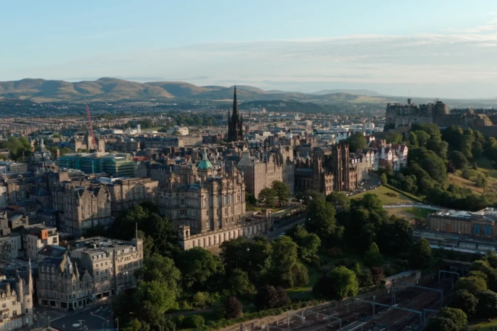 Aerial view of Edinburgh showcasing historic architecture, lush greenery, and the backdrop of rolling hills under a clear sky.