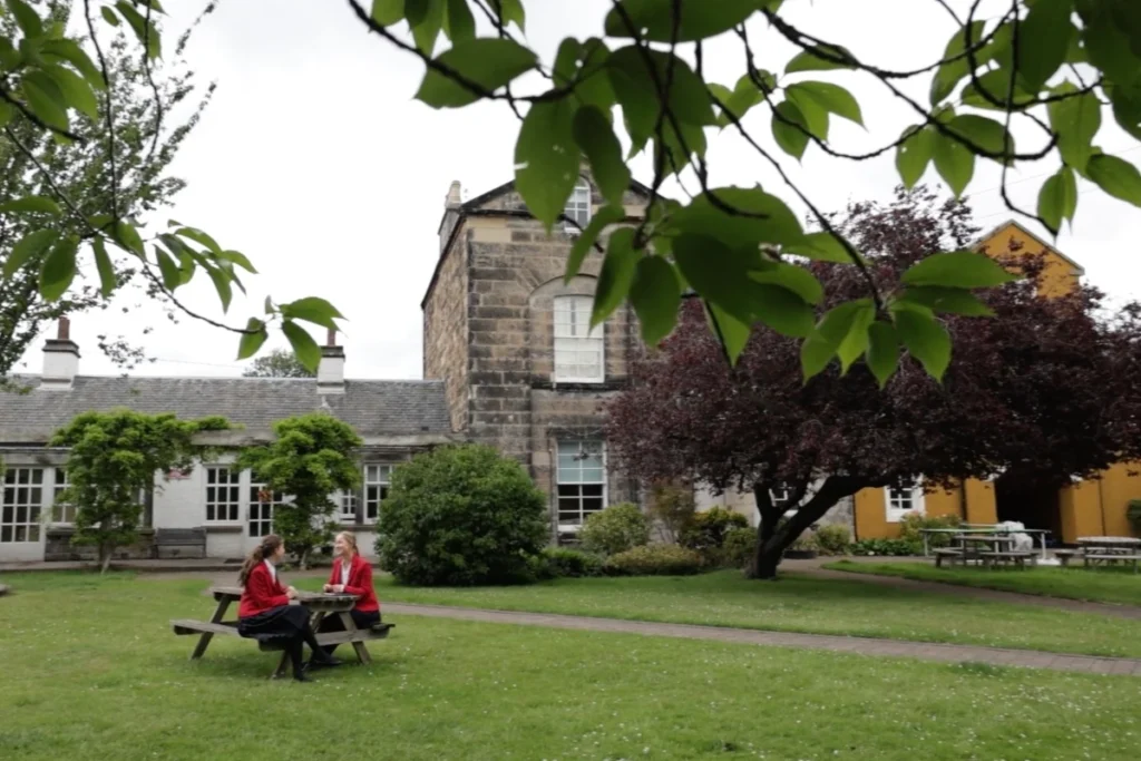 Two Loretto students in their red blazers sitting at a picnic table in a lush garden, surrounded by trees and the historic school building in the background.