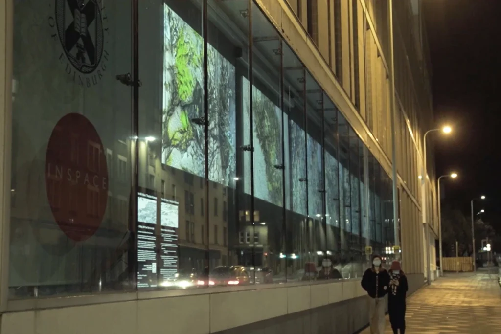 Two people walk past a modern building at night, illuminated by colourful projections on its glass facade in Edinburgh.