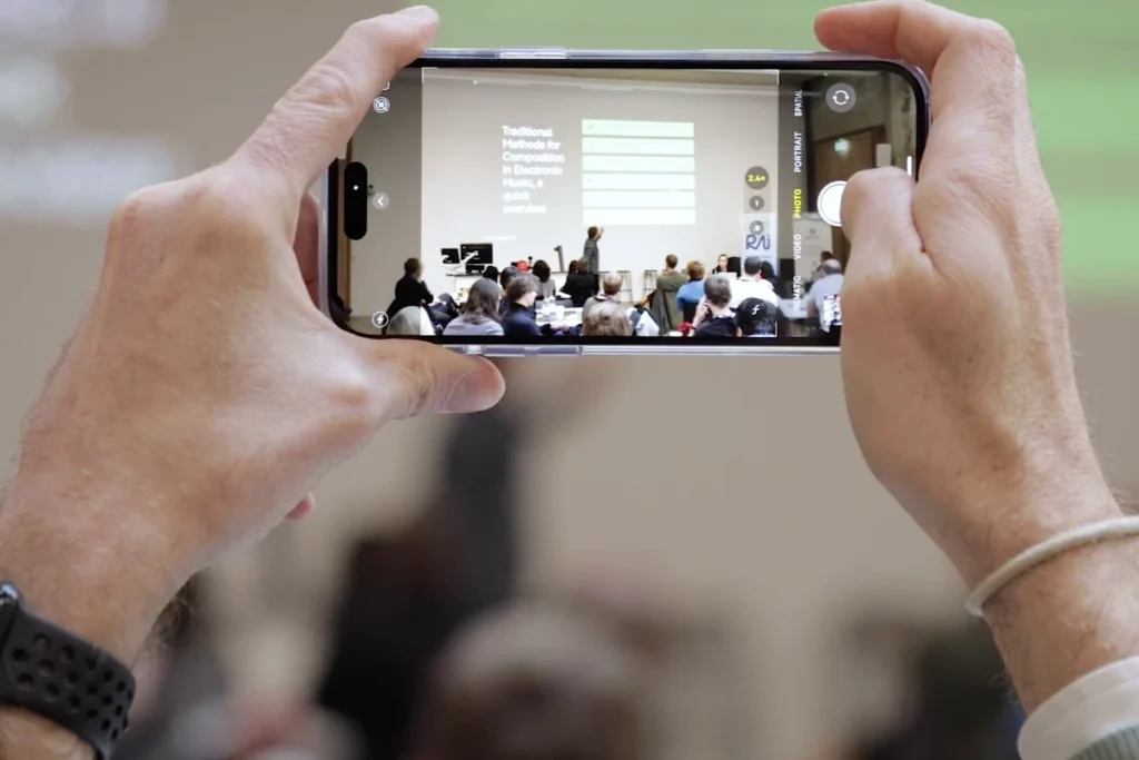A person capturing a presentation on their smartphone during a workshop on electronic music composition methods.