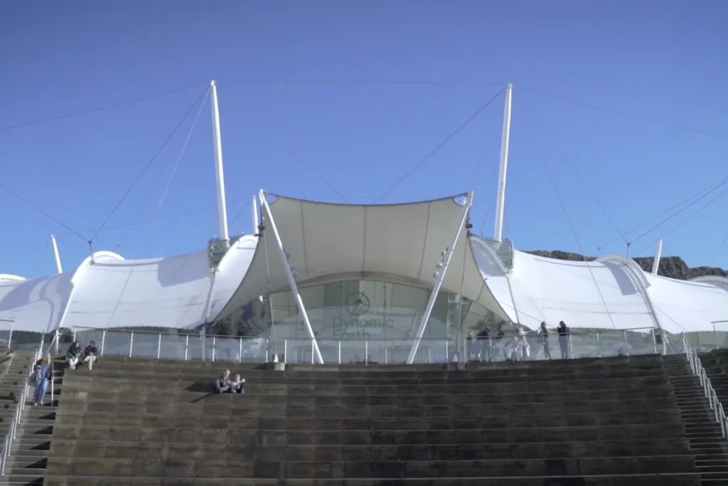 Edinburgh Dynamic Earth building with modern architecture, featuring a large white canopy and visitors on the steps under a clear blue sky.