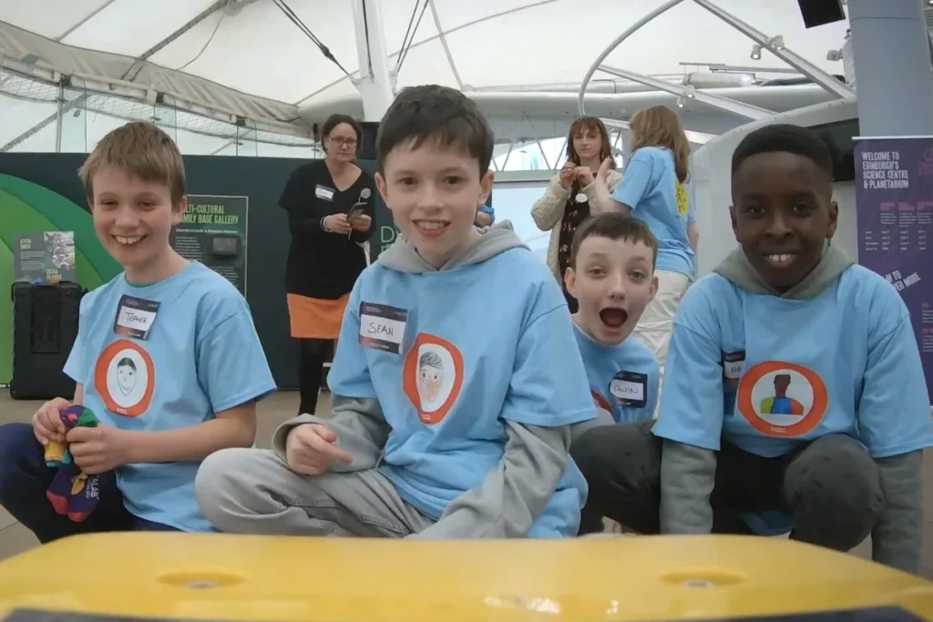 Four children wearing blue shirts smile and pose for the camera at the Scottish AI Summit Promotional event, showcasing joy and excitement.