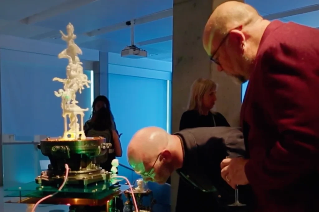 Two men with glasses in their hands look out over a complex, white sculpture on a green pedestal in an exhibition room with other people in the background